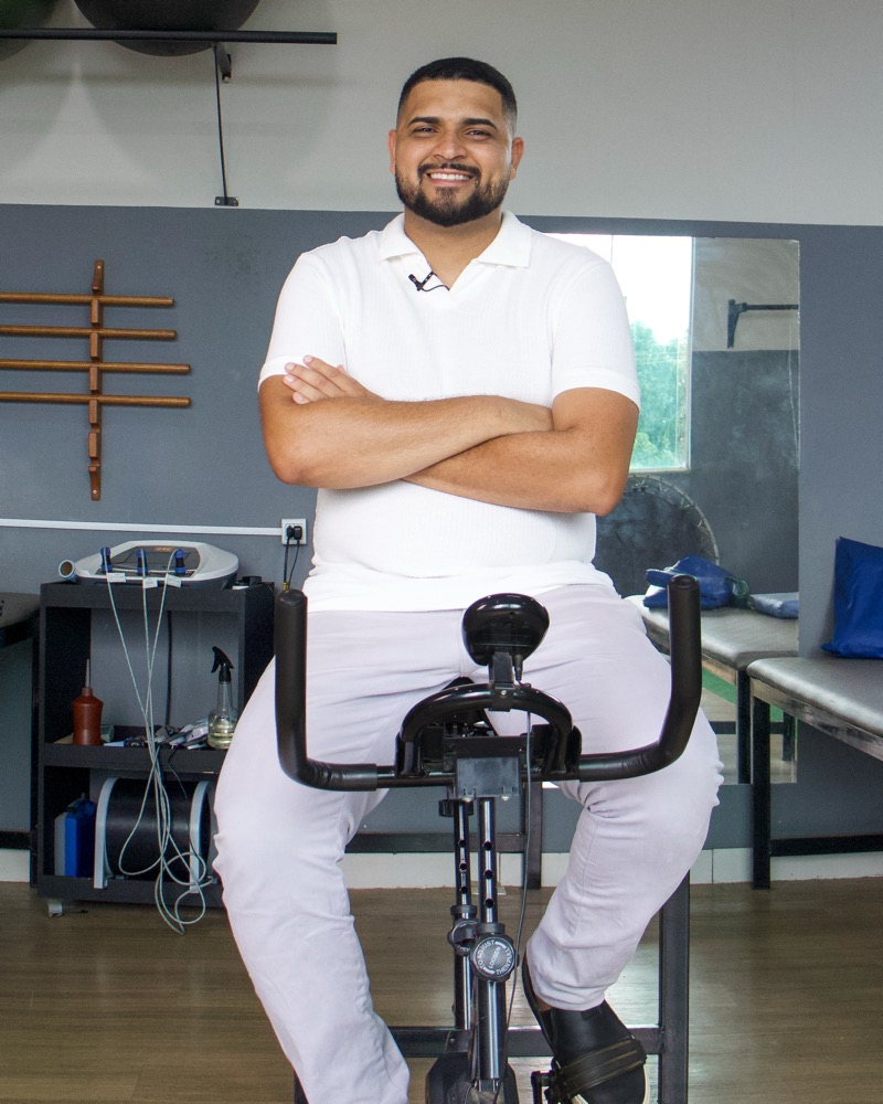 Jefferson Lima sorrindo na área de reabilitação da clínica, com bicicleta e aparelhos